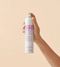 Hand holding a 'Make Me Shine Spray Gloss' by Eleven on a beige background - Hair Care Canada