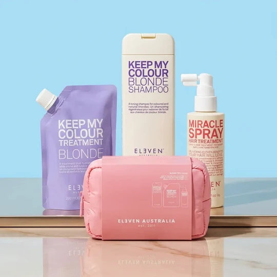 A set of Eleven Australia hair color care products arranged on a reflective surface against a light blue background, including a pink zip pouch, a purple treatment pouch, a white shampoo bottle, and a beige hair spray bottle.