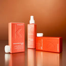 Three Kevlin Murphy Everlasting Colour Care products in a matching coral-orange color, arranged on a reflective brown surface against a warm brown background. The set includes a wash bottle with a white cap lying beside it, a spray bottle with a white spray top, and a rectangular box with a white lid.