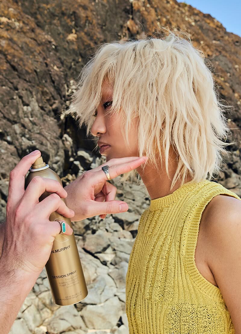Person applying hair spray to another person's hair outdoors with a rocky background - Hair Care Canaa