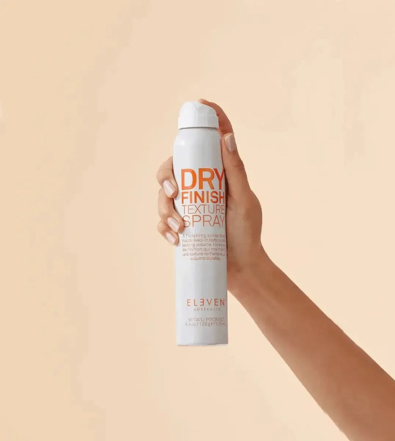 Hand holding a can of 'Dry Finish Texture Spray' against a beige background - Hair Care Canada