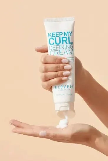 Hand holding a tube of 'Keep My Curl Defining Cream' by Eleven on a beige background - Hair Care Canada