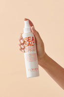 Hand holding a bottle of 'Sea Salt Texture Spray' by Eleven Australia against a beige background - hair Care Canada