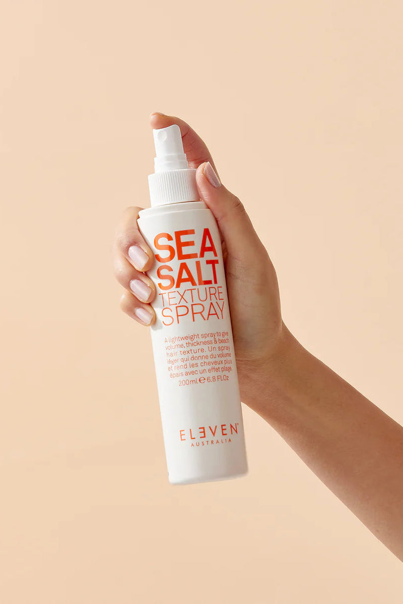 Hand holding a bottle of 'Sea Salt Texture Spray' by Eleven Australia against a beige background - hair Care Canada