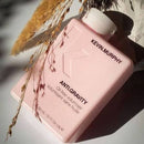 A pink rectangular bottle of Kevin Murphy Anti-Gravity Oil Free Volumiser with a white cap, styled with dried grass stems on a white surface under direct sunlight.