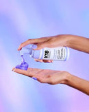 A person's hands holding a bottle of K18 hair color and dispensing purple hair dye onto a hand, set against a soft purple and blue gradient background.