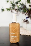 A bottle of Kryolan Shimmer Shine Shined Hair Spray with a white spray top and clear cap, filled with a shimmering golden-brown liquid, set on a dark surface with a blurred floral background.