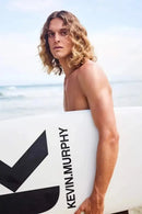 Man holding a surfboard with 'Kevin Murphy' branding on a beach Hair Care Canada