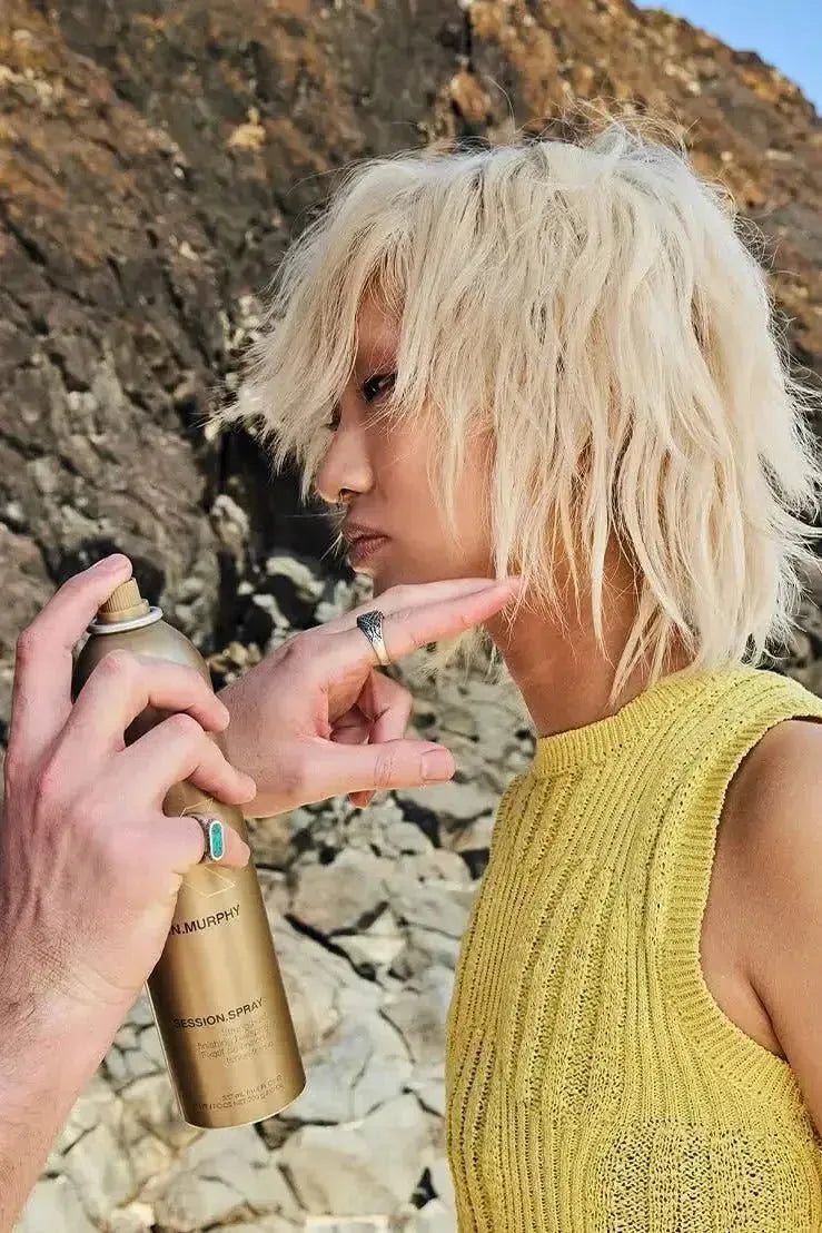 Person applying hair spray to another person's hair outdoors with a rocky background - Hair Care Canaa